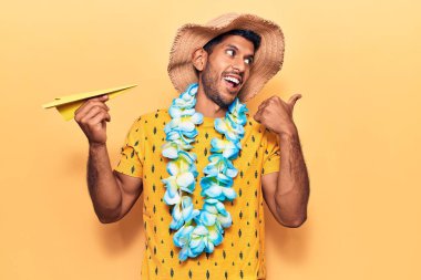 Yaz şapkası takan genç latin adam ve kağıt uçak tutan Hawaii zambağı başparmağını yan tarafa uzatıp ağzı açık mutlu bir şekilde gülümsüyor. 