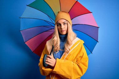 Yağmurlu hava için yağmurluk giyen genç ve güzel sarışın bir kadın renkli bir şemsiye tutuyor ve akıllı bir yüz ifadesiyle ciddi ciddi düşünüyor.
