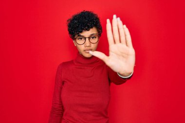 Young beautiful african american afro woman wearing turtleneck sweater and glasses doing stop sing with palm of the hand. Warning expression with negative and serious gesture on the face. clipart