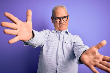 Orta yaşlı, beyaz, çizgili gömlek ve mor arka planda gözlük takan yakışıklı adam kameraya sarılıp gülümsüyor. Mutluluğu kucaklayan neşeli bir ifade..