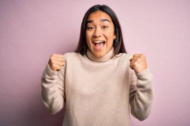 Genç ve güzel Asyalı kadın pembe arka plan üzerine balıkçı yaka kazak giyip kollarını kaldırarak ve açık gözlerle başarıyı kutluyor. Kazanan konsept.