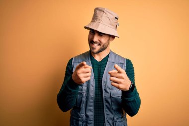 Tatilde sakallı, sarı arka planda kaşif şapkası takan yakışıklı turist mutlu ve komik yüzlü parmaklarını kameraya doğrultuyor. İyi enerji ve titreşimler.