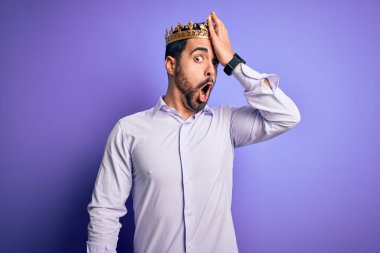 Young handsome man with beard wearing golden crown of king over purple background surprised with hand on head for mistake, remember error. Forgot, bad memory concept. clipart