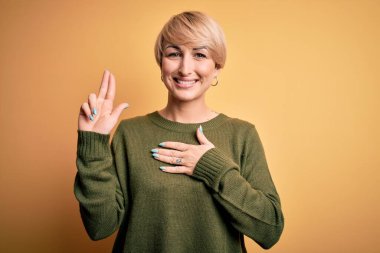 Modern kısa saçlı genç bir kadın sarı arka planda rahat süveter giyiyor gülümsüyor, elleri göğsünde ve parmakları havada küfür ediyor, sadakat yemini ediyor.