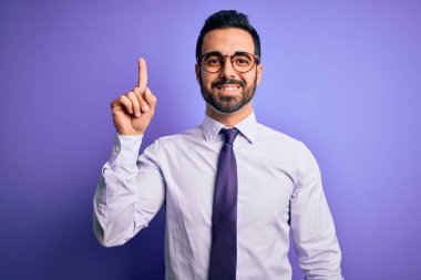 Sakallı, günlük kravat takan ve mor arka planda gözlüklü yakışıklı bir iş adamı başarılı bir fikri işaret ediyor. Heyecanlı ve mutlu. Bir numara..