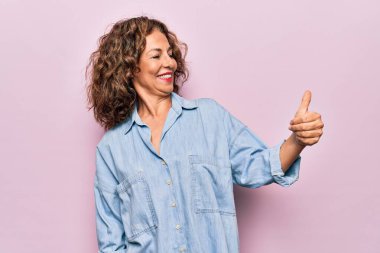 Orta yaşlı, güzel bir kadın pembe arka plan üzerinde sıradan bir kot gömlek giyiyor gururlu görünüyor, gülümsüyor yan tarafa başparmak işareti yapıyor.