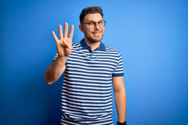 Mavi gözlü, gözlüklü mavi çizgili tişörtlü, mavi arka planda dört numarayı gösteren ve mutlu bir şekilde gülümseyen genç bir adam..