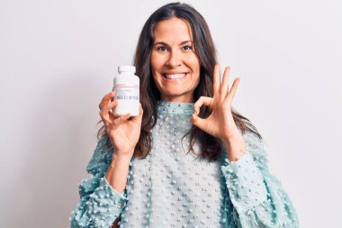 Genç, güzel esmer kadın ilaç kavanozunu izole edilmiş beyaz arka planda tutuyor. Parmaklarıyla işaret veriyor, gülümsüyor. Mükemmel bir sembol.