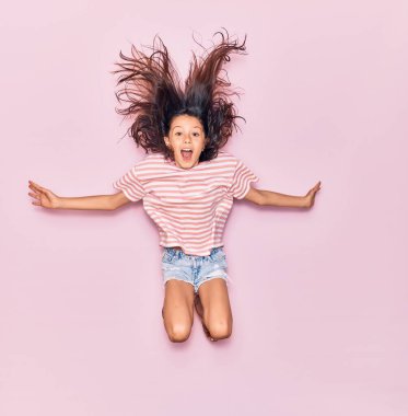 Sevimli İspanyol kız günlük kıyafetler giyip mutlu gülümsüyor. Yüzünde ve kollarında gülücüklerle pembe bir arka planda atlamak.