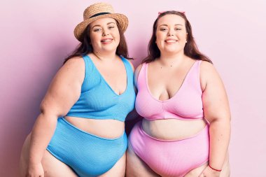 Bikini giyen genç ve cüsseli ikizler pozitif ve mutlu görünüyorlar. Ayakta duruyorlar ve gülümsüyorlar. 