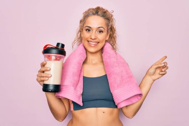 Güzel, sarışın, spor yapan, havluyla protein içeceği sallayan, eli ve parmağı kenarda mutlu gülümseyen bir kadın.