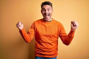 Sarı arka plan üzerinde duran turuncu süveter giyen genç yakışıklı adam kollarını kaldırarak ve açık gözlerle başarıyı kutluyor ve şaşırıyor. Kazanan konsept.