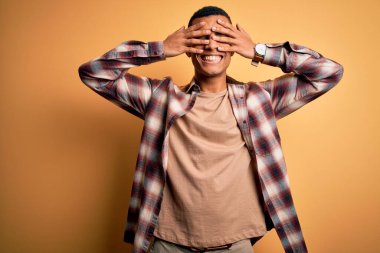 Genç, yakışıklı, Afro-Amerikan bir adam, günlük gömleğini giymiş, sarı arka planda duruyor, gözleri güler yüzlü ve komik bir şekilde kapatıyor. Kör kavram.