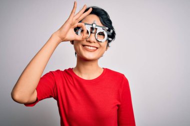 Optometri gözlüğü takan genç, güzel Asyalı kız izole edilmiş beyaz arka planda duruyor. El hareketleriyle gülümsüyor, gözleriyle mutlu yüzüyle parmaklarının arasından bakıyor..