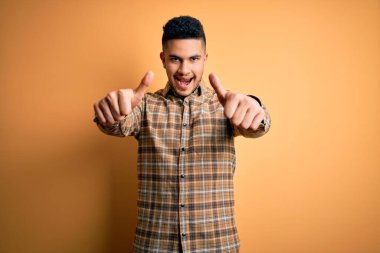 Genç yakışıklı adam izole sarı arka plan üzerinde duran rahat gömlek giyen el ile olumlu jest yapıyor onaylayan, gülümseyerek kadar başparmak ve başarı için mutlu. Kazanan jest.