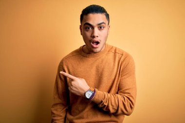 İzole edilmiş sarı arka planda dikilen sıradan bir süveter giyen genç Brezilyalı adam parmağını yana doğru göstererek şaşırdı, ağzı açık şaşkın bir ifade takındı..