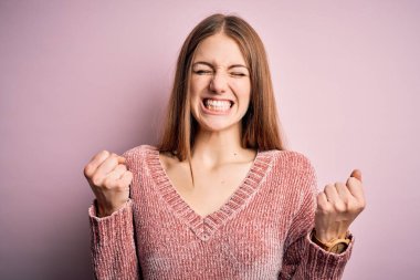 İzole edilmiş pembe arka planda rahat bir süveter giyen genç ve güzel bir kadın. Kolları havada ve gözleri kapalı zaferi kutlarken gülümsüyor. Kazanan konsept.