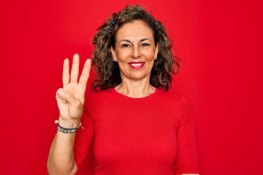 Orta yaşlı, esmer, sıradan tişörtlü bir kadın kırmızı arka planda dikiliyor ve üç numarayı gösterirken kendinden emin ve mutlu bir şekilde gülümsüyor..