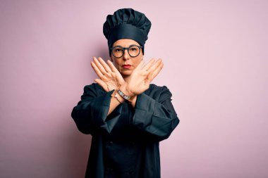Orta yaşlı esmer aşçı kadın, aşçı üniforması giyiyor ve şapka takıyor. Pembe arka plan üzerinde. Reddedilme ifadesi.