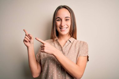 İzole edilmiş beyaz arka planda dikilip gülümseyen ve kamerayı iki eliyle ve parmaklarıyla gösteren genç ve güzel bir kadın..