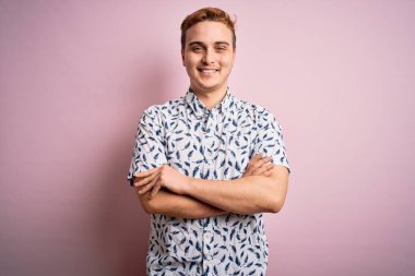 Genç, yakışıklı, yaz gömleği giyen, pembe arka planda duran, kollarını kavuşturup kameraya gülümseyen bir adam. Pozitif kişi.