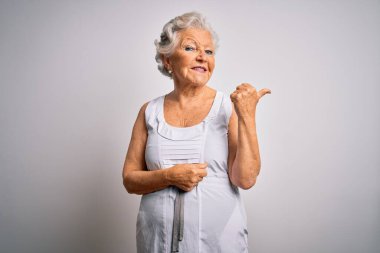 Son sınıf güzeli, gri saçlı, beyaz arka planda rahat bir yaz elbisesi giyen mutlu yüzüyle gülümseyen ve baş parmağıyla yüzünü gösteren bir kadın..