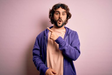 Genç, yakışıklı, sakallı, pembe arka planda sıradan bir kazak giyen, ağzı açık, parmaklarını yana doğru gösteren, ağzı açık, şaşırmış bir adam..