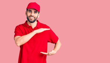 Sakallı, teslimat üniforması giyen, elleri büyük ve büyük boy işareti gösteren genç yakışıklı bir adam. Ölçü sembolü. Kameraya bakıp gülümsüyor. ölçüm kavramı. 