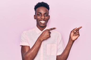 Genç Afro-Amerikalı sıradan kıyafetler giyen, gülümseyen ve kameraya bakan iki elini ve parmağını yana doğru gösteren bir adam.. 