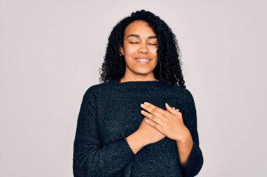 Beyaz arka plan üzerinde sıradan bir süveter giyen, elleri göğsünde, gözleri kapalı ve yüzünde minnet dolu bir jestle gülümseyen Afro-Amerikan kıvırcık kadın. Sağlık kavramı.