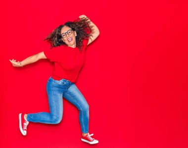 Orta yaşlı, güzel İspanyol bir kadın günlük kıyafetler ve mutlu bir gülümseme takınıyor. İzole edilmiş kırmızı arka planda yüzünde gülümsemeyle zıplıyor