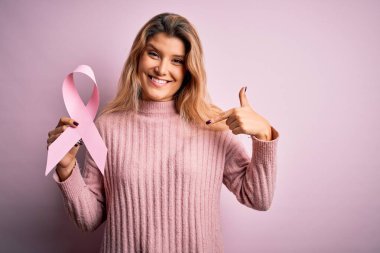 Genç ve güzel sarışın bir kadın kanseri desteklemek istiyor elinde pembe kurdele sembolü ve kendini gösteren sürpriz bir yüz var.