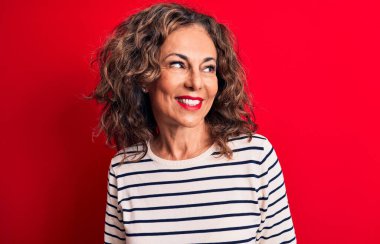 Orta yaşlı, esmer, çizgili tişört giyen güzel bir kadın kırmızı arka planda duruyor. Yan tarafa bakıyor, rahat bir poz veriyor ve kendine güvenen bir gülümseme sergiliyor..