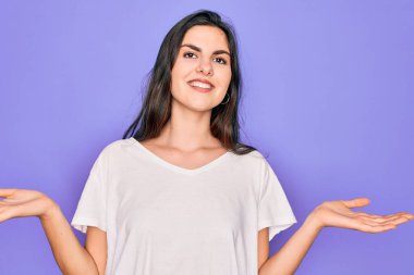 Genç, güzel esmer kadın, mor arka plan üzerine günlük beyaz tişört giyip gülümsüyor. Her iki elinde açık avuçlar, sunum ve reklam karşılaştırma ve denge gösteriyor.