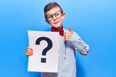 Sevimli sarışın çocuk inek papyonu takıyor ve gözlük takıyor. Elinde soru işareti var. Mutlu ve olumlu gülümsüyor. 