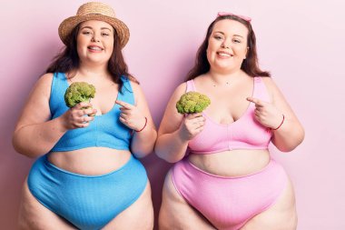 Bikini giyen, brokoli tutan ve mutlu gülümseyen genç ve büyük beden ikizler. 