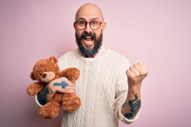 Sakallı ve dövmeli yakışıklı kel adam pembe arka planda oyuncak ayı tutuyor gururla bağırıyor ve zaferi ve başarıyı kutluyor.