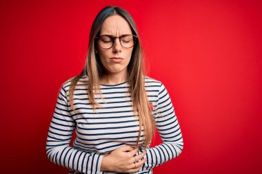 Mavi gözlü, genç, sarışın bir kadın. Kırmızı arka planda duruyor. Midesinde hazımsızlık var. Ache kavramı.