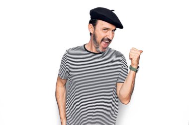 Orta yaşlı, çizgili tişört giyen ve beyaz arka planda Fransız beresi takan yakışıklı bir adam baş parmağını yana uzatıp ağzı açık gülümsüyor.