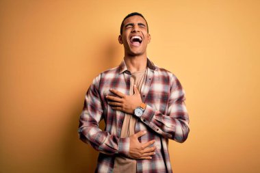 Genç, yakışıklı, Afro-Amerikalı bir adam sarı arka planda dikilmiş gülümsüyor ve kahkahalarla gülüyor..