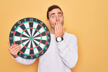 Mavi gözlü genç adam, izole edilmiş sarı arka planda dart tahtasını tutuyor. Ağzı kapalı, utancından ağzı açık, korkusunun dışa vurumu, sessizlikten korkması, gizli konsept.