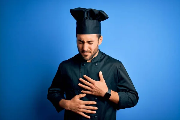 Young handsome chef man with beard wearing cooker uniform and hat over blue background with hand on stomach because indigestion, painful illness feeling unwell. Ache concept.