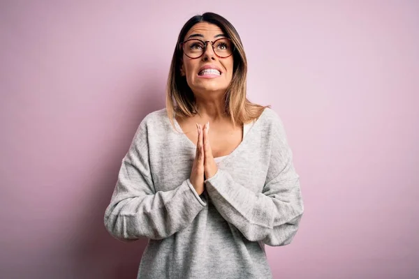 Young beautiful brunette woman wearing casual sweater and glasses over pink background begging and praying with hands together with hope expression on face very emotional and worried. Begging.