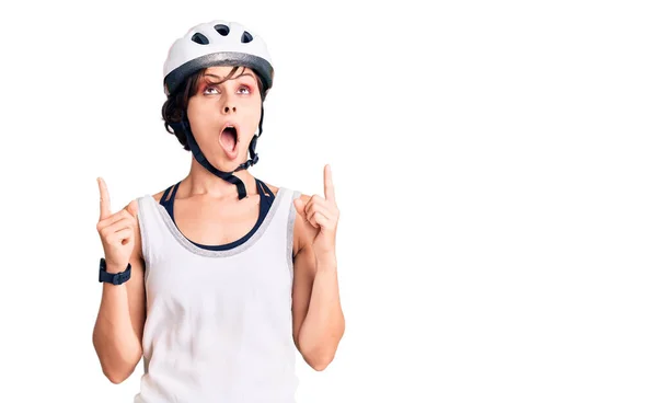 Beautiful young woman with short hair wearing bike helmet amazed and surprised looking up and pointing with fingers and raised arms.