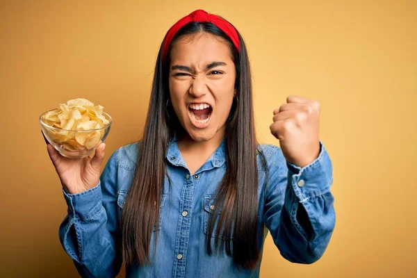 年轻美丽的亚洲女人拿着盛有土豆片的碗 在孤立的黄色背景下 怒气冲冲地大叫着 发疯了 高喊着 怒气冲冲 — 图库照片