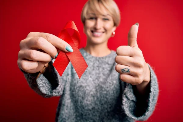 Young blonde woman with short hair holding hiv and aids awareness red ribbon happy with big smile doing ok sign, thumb up with fingers, excellent sign