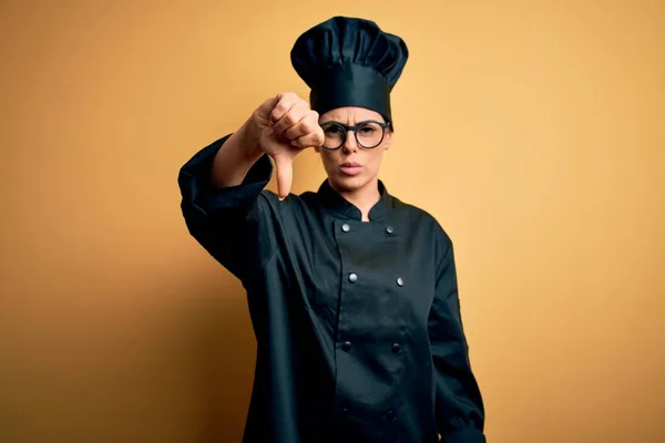 Young beautiful brunette chef woman wearing cooker uniform and hat over yellow background looking unhappy and angry showing rejection and negative with thumbs down gesture. Bad expression.