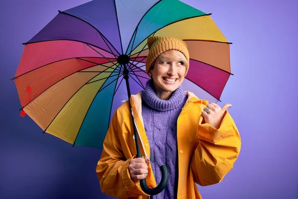 年轻的金发女子 身穿防水雨衣 头戴彩色雨伞 手指朝天 笑容满面 笑容满面 — 图库照片
