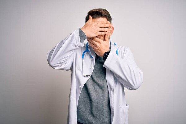 Young doctor man with blue eyes wearing medical coat and stethoscope over isolated background Covering eyes and mouth with hands, surprised and shocked. Hiding emotion