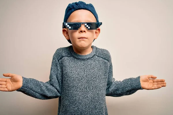 Young little caucasian kid wearing internet meme thug life glasses over isolated background clueless and confused expression with arms and hands raised. Doubt concept.